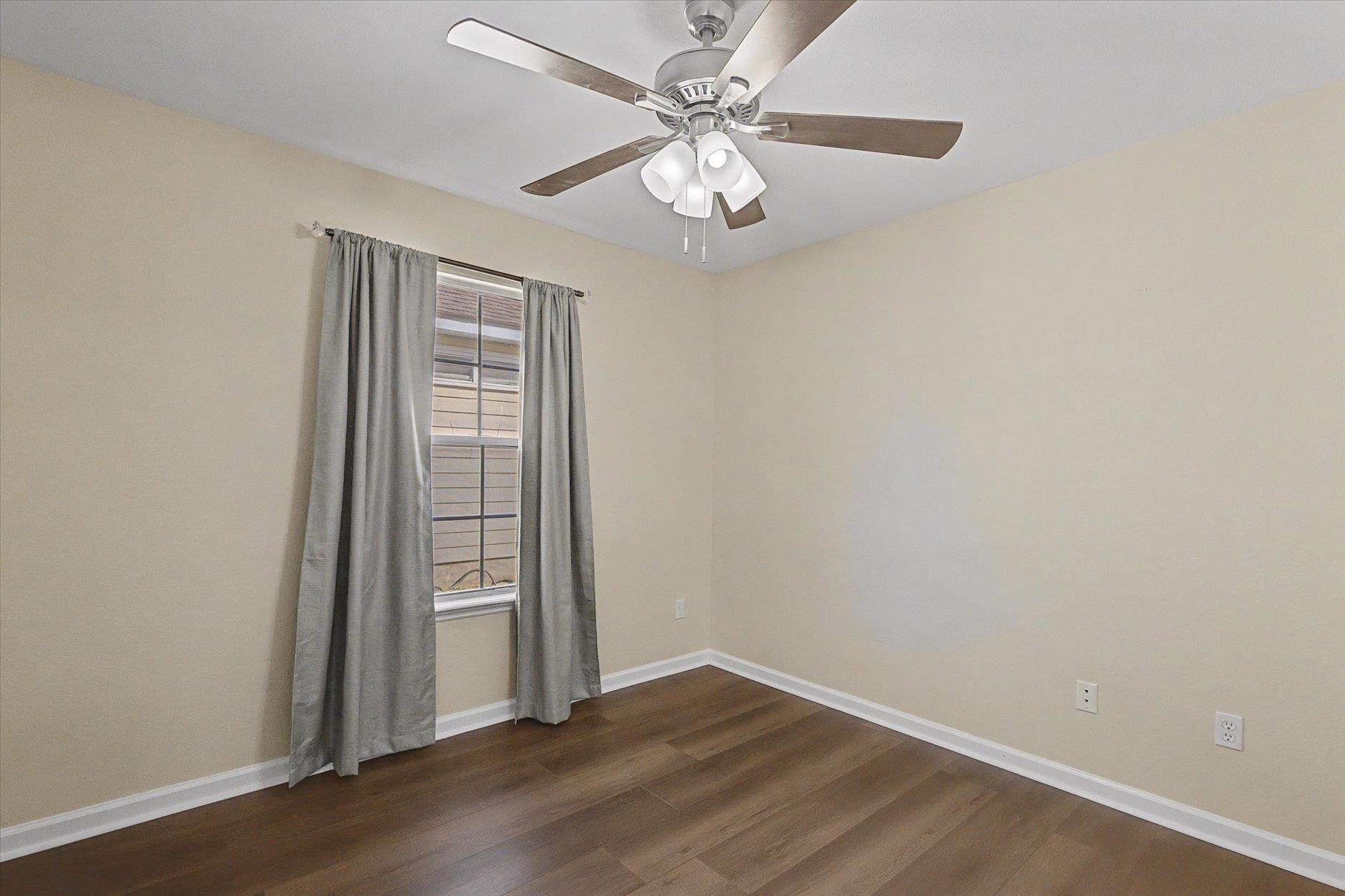 Empty room with wooden floor, ceiling fan, and window with gray curtains.