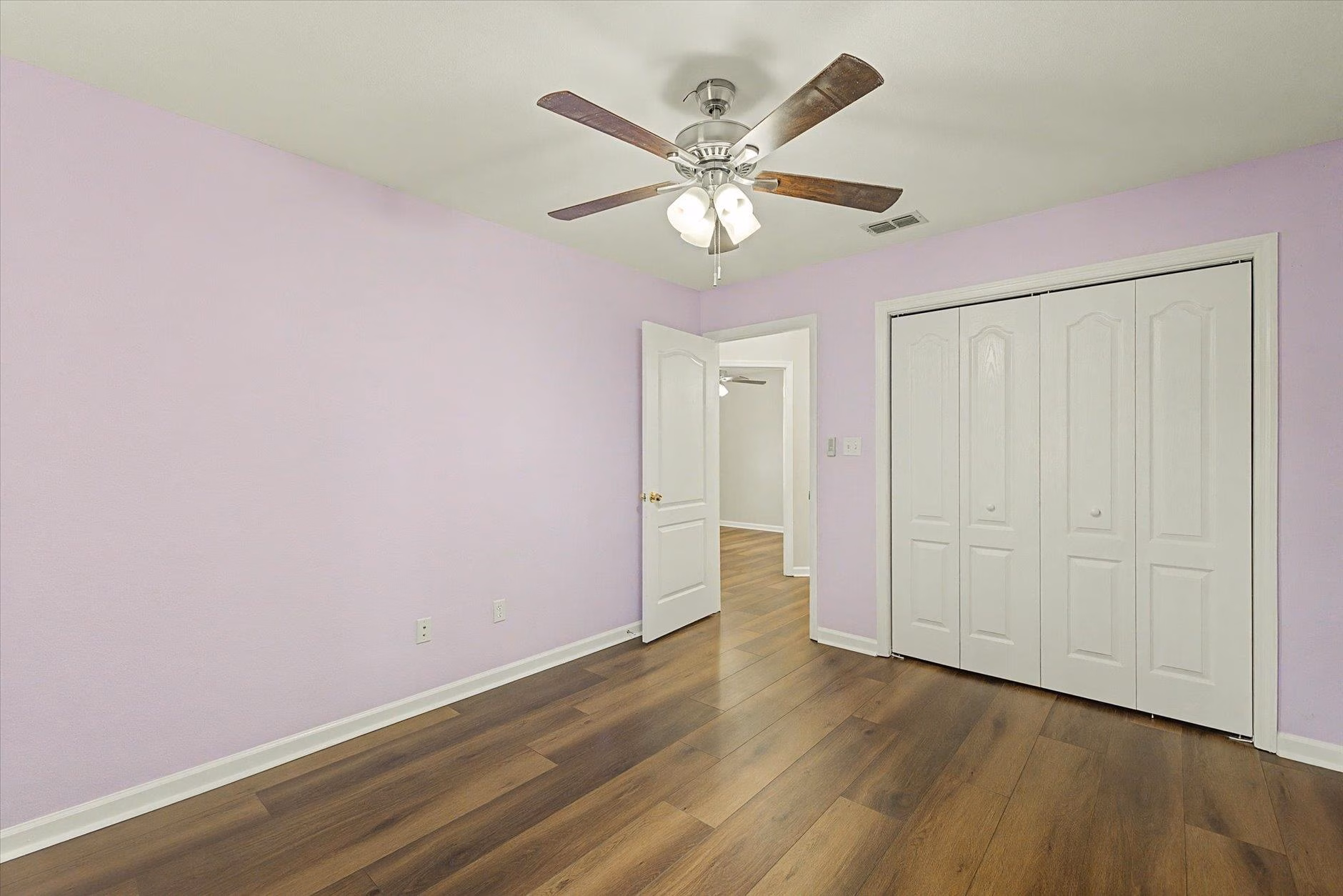 Empty room with lavender walls, wooden floor, ceiling fan, and closet doors.
